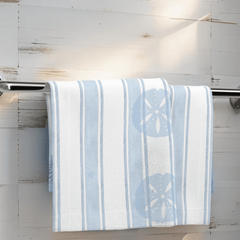 Light blue coastal face towel with sand dollar design hanging on rack in beach-themed bathroom.